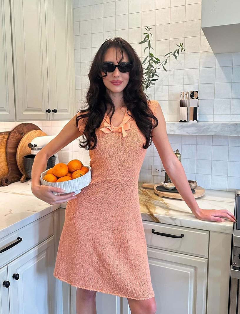 Sydney Carlson poses in kitchen, wearing a coral dress, holding a bowl of oranges