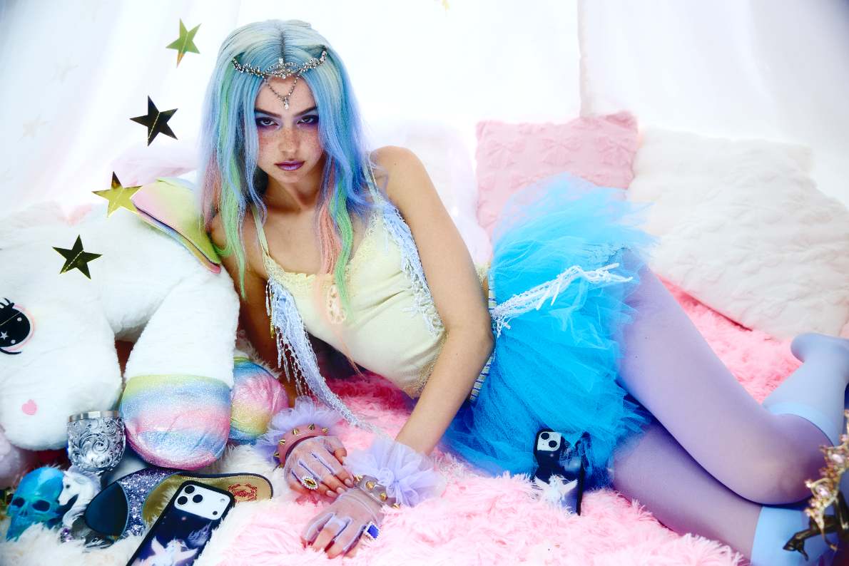 Mazzy Joya lays on a pink fluffy bed spread wearing a blue tutu and stockings, yellow tank top, and a blue and green wig