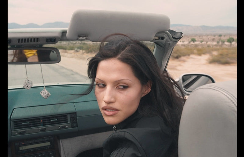 Gabbriette sits passenger in a teal convertible car, while looking back  
