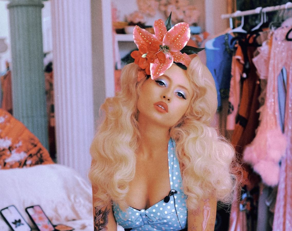 Remy Bond wears a large orange flower hair accessory and blue polka dot top
