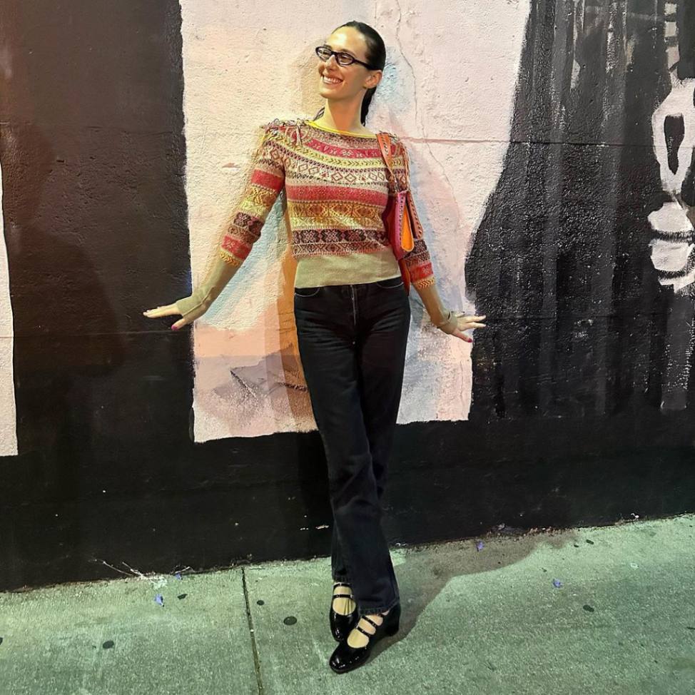 Devon Lee Carlson poses against a wall in a striped sweater and black denim jeans