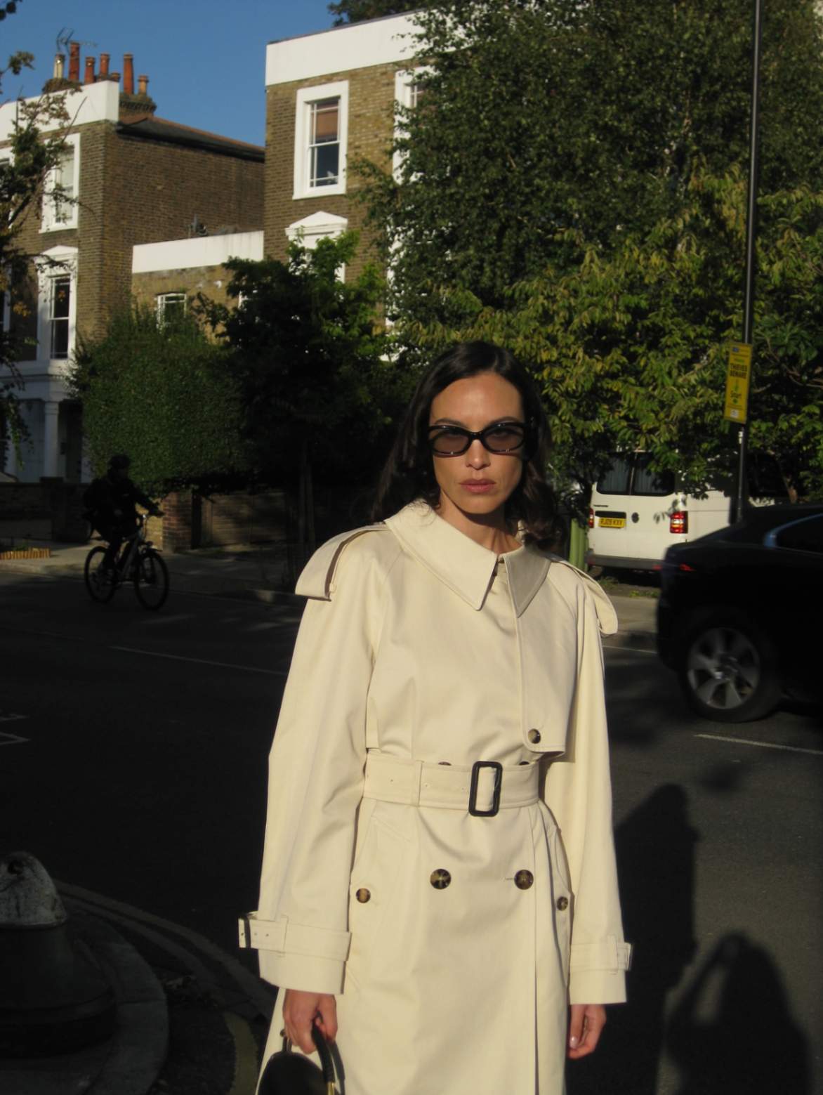 A woman poses outside in a white trench coat and black sunglasses