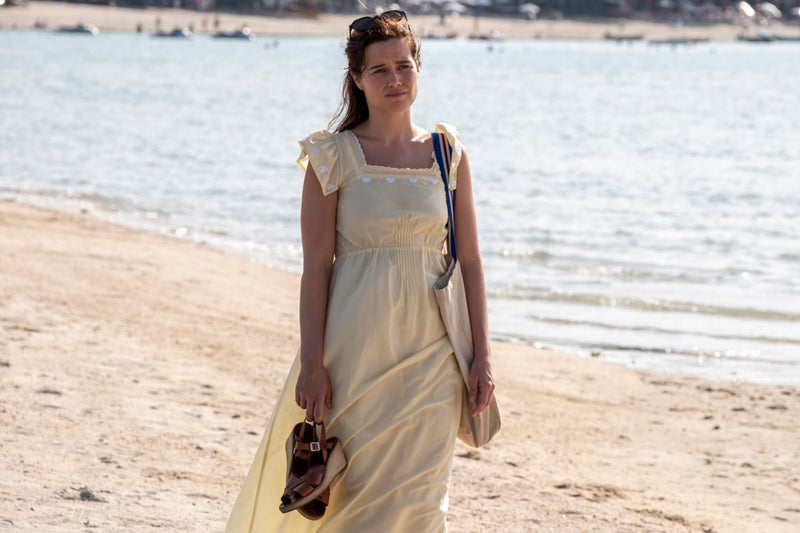 A woman stands on sand on the beach in a long yellow dress and holds her sandals